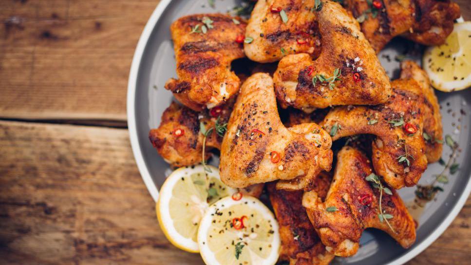 A plate of seasoned chicken wings, served with chilli garnish and wedges of lemon. The grey plate they are served on is sitting on a wooden table. 