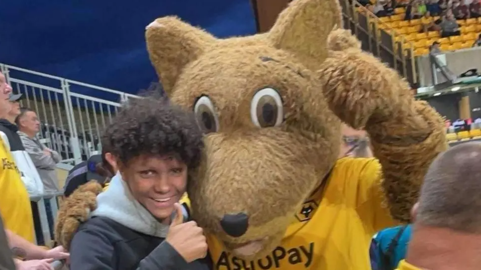 A young boy with a thick mop of curly black hair stands next to a football mascot, a wolf wearing a yellow Wolves shirt, in a football stadium. Both are giving the thumbs-up to the camera