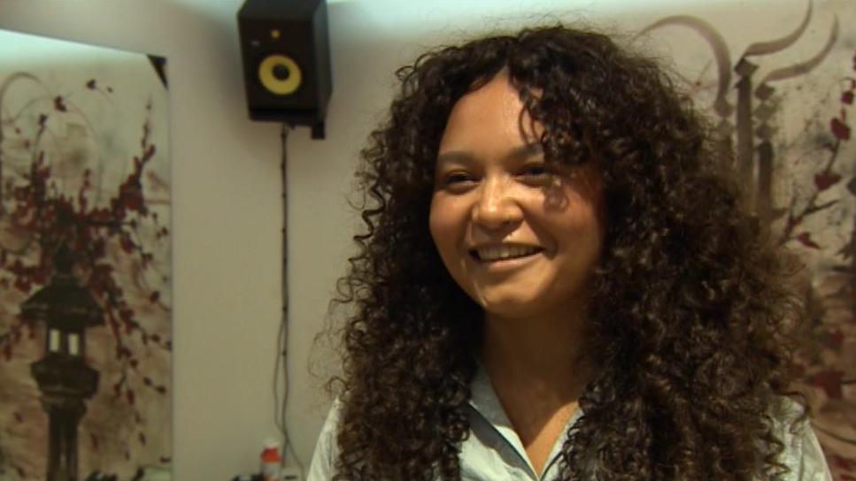 A woman with long dark curly hair and wearing a blue shirt looks at the camera. Behind her there is artwork on the wall and a speaker.  