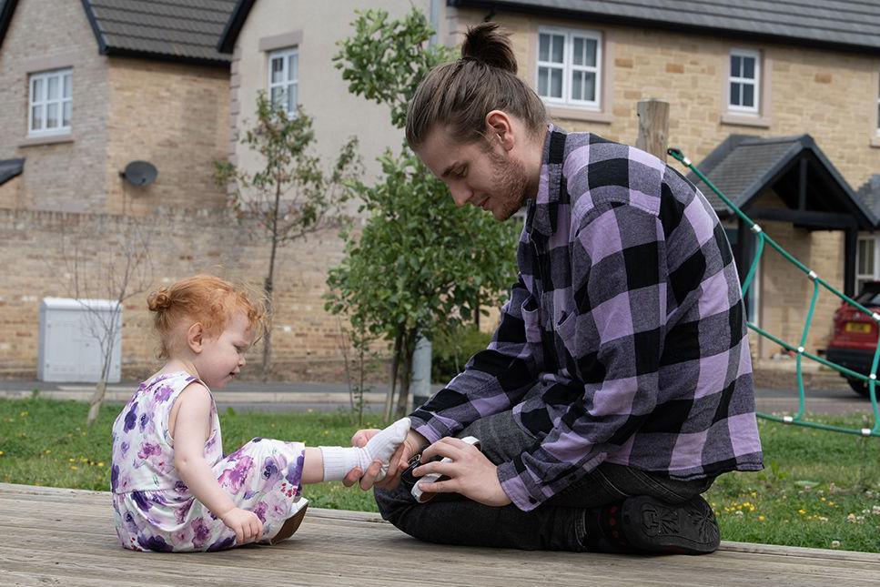 James, who's wearing a light and dark purple checked shirt and has his hair in a topknot, is holding his daughter's white-socked foot, ready to put on a shoe. They're both sitting on decking and she has red hair and is wearing a white and purple floral dress.