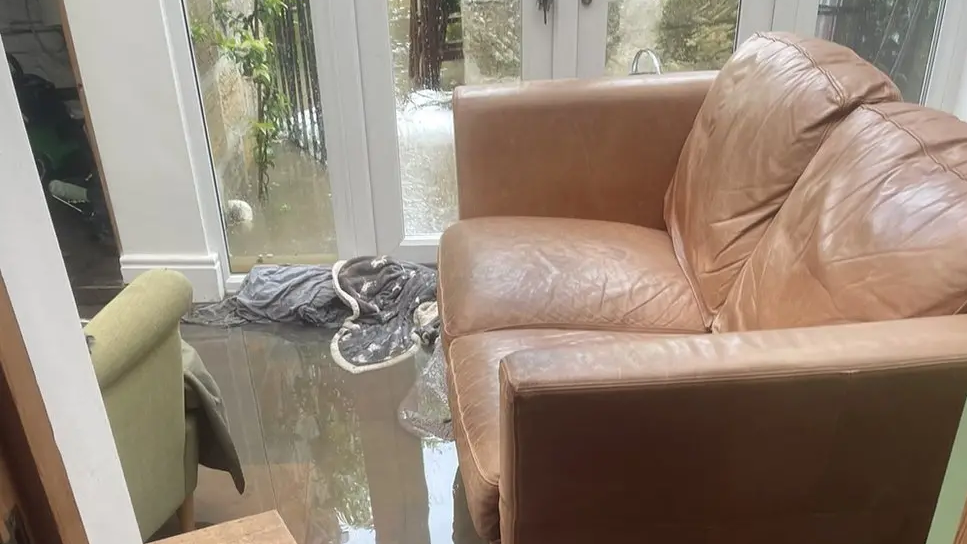 Brown leather sofa standing in grey flood water a few inches deep in a lounge. There are with soaked towels next to it beside a window. 