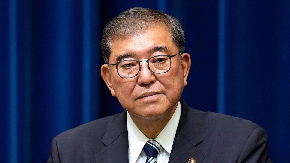 Japan's Prime Minister Shigeru Ishiba wearing a dark suit stands in front of blue curtains