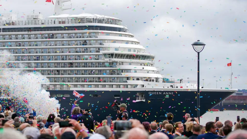 A wide shot of the Queen Anne cruise ship arriving into the Mersey. It is an overcast day, but hundreds of spectators are celebrating with colourful confetti.