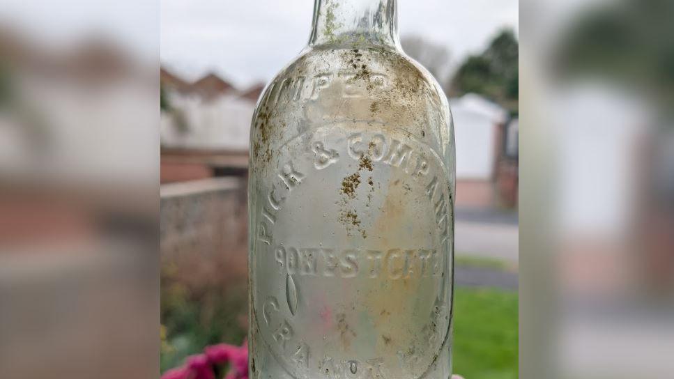 A clear thin-necked bottle with mud on it being held up. An inscription on the front reads "Imperial, Pick & Company, Westgate, Grantham."