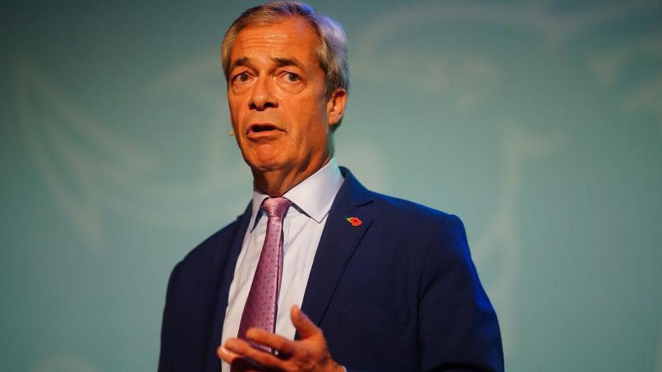 Nigel Farage wearing a blue suit, with a light blue shirt underneath and a pink tie. He is about to speak and has a mic on his face.