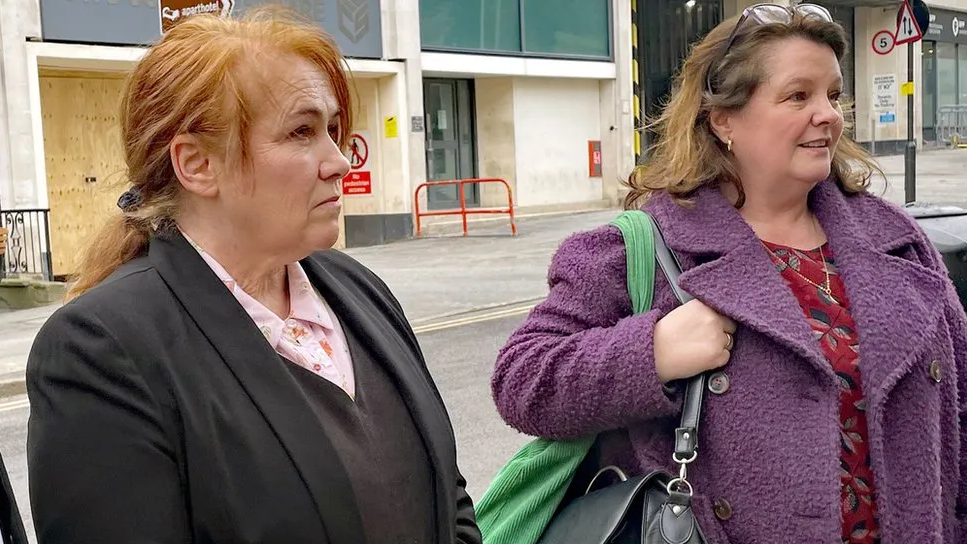 Two women stand on a street, looking to the right at a person off-camera. The woman on the left, Joanne Billington, has ginger hair tied back, and is wearing a pink shirt, black jumper, and black jacket. The woman on the right wears a red top and a purple coat.