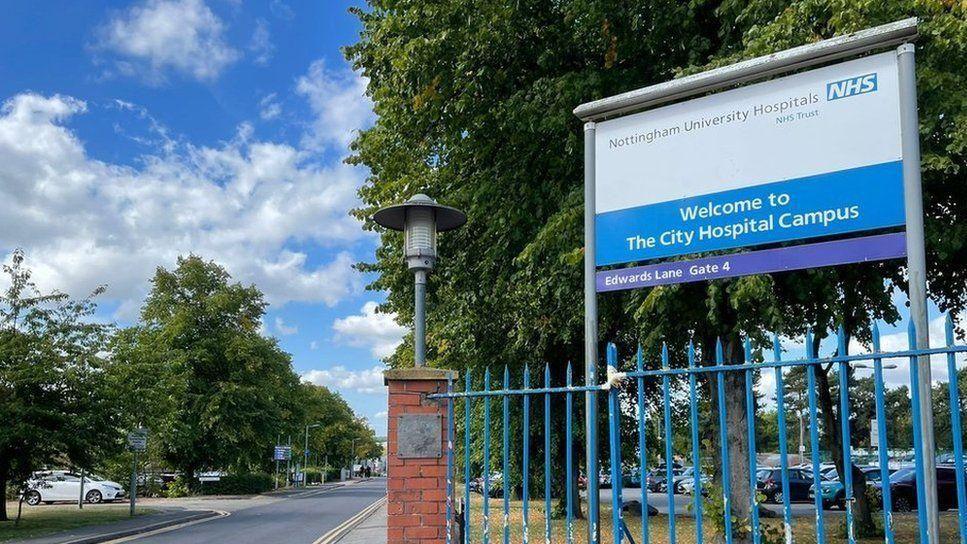 The entrance to the City Hospital Campus, with a tree-lined road stretching into the distance on the left and blue metal railings and a sign for the hospital to the right

