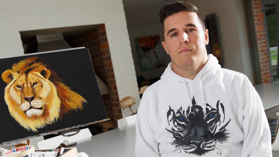 Henry Fraser in sitting on the right. He has brown hair and is wearing a white hoodie with a black and white image of a tiger on it. He is sitting next to one of his paintings of a male lion.