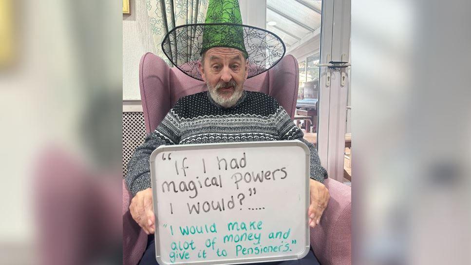 An man wearing a grey detailed jumper sitting in a pink chair, wearing a witches hat and holding up a sign that says "if I had magical powers I would?" with the answer "I would make a lot of money and give it to pensioners".