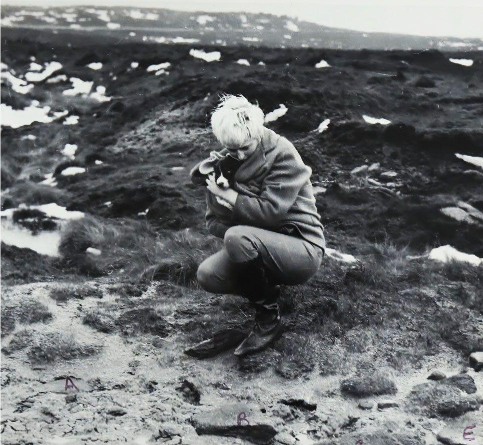 A black and white photograph shows Myra Hindley crouching on a rocky, grassy terrain. She is wearing a coat and boots, and is holding a small dog close to her chest. Snow patches are visible in the background.