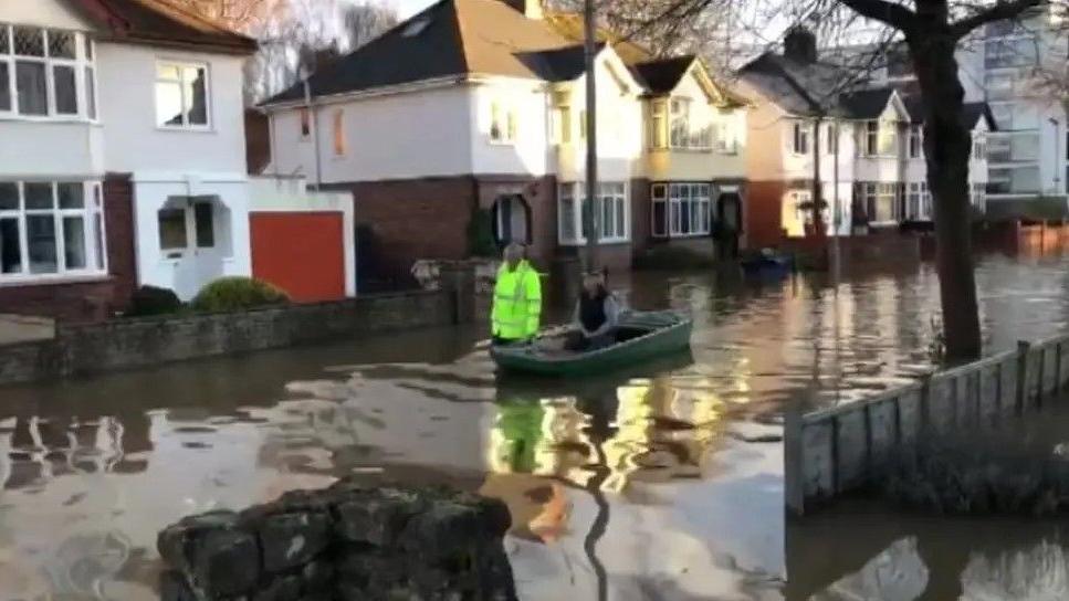 A row of semidetached houses can be seen amid a flooded road with a boat in the middle where someone wearing a high visibility jacket is stood in ithe water 