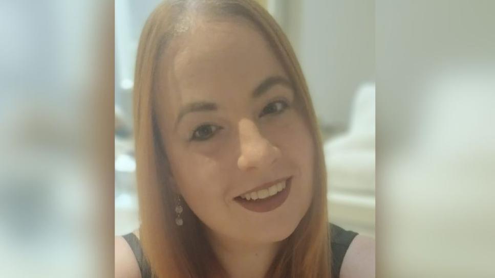 The photo shows a woman smiling with ginger hair and wearing a black top