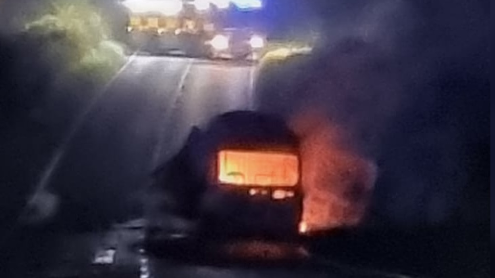 A blurry picture with a fire engine with its lights on at the top of the road, with a van clearly on fire with what appears to be flames in its cabin