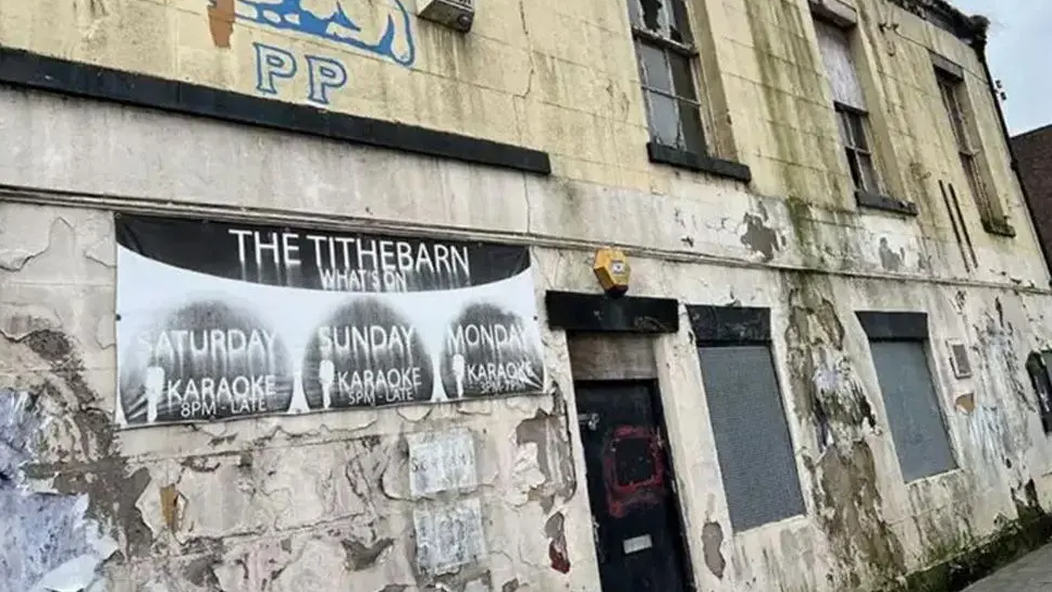 A close-up of the Tithebarn pub. It is an old cream-coloured structure with peeling paint. There is a faded sign describing what events were going on at the pub.