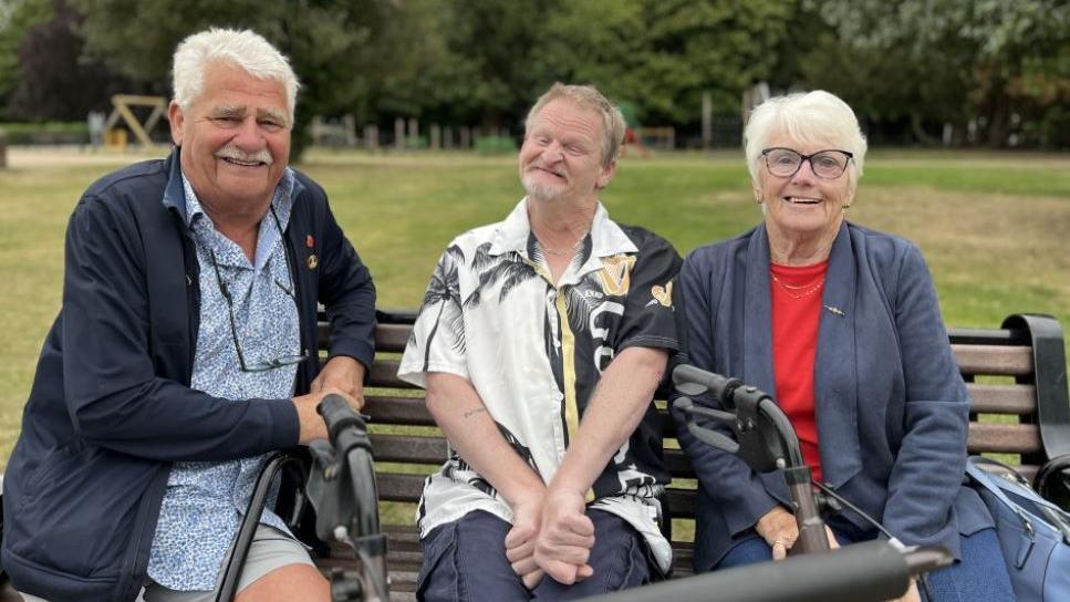 A man with short grey hair and a moustache in a blue shirt and jacket leaning next to a park bench. On the bench is a man smiling in a black and white shirt with a woman sat next to him with glasses and short blonde hair.