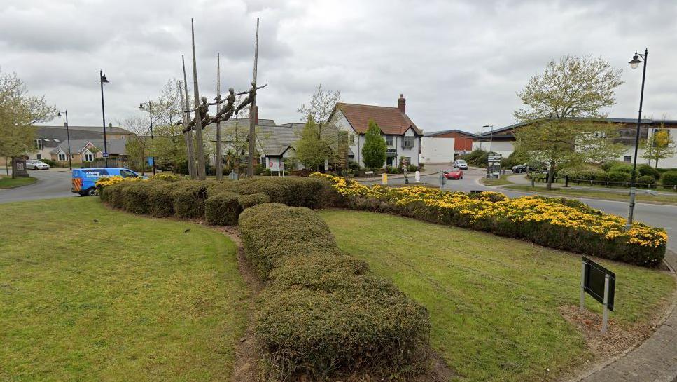 A landscaped roundabout in Ravenswood, Ipswich, with shrubs, gorse and a sculpture of six flying metal human figures, with their arms outstretched. Cars and buildings are visible behind the roundabout