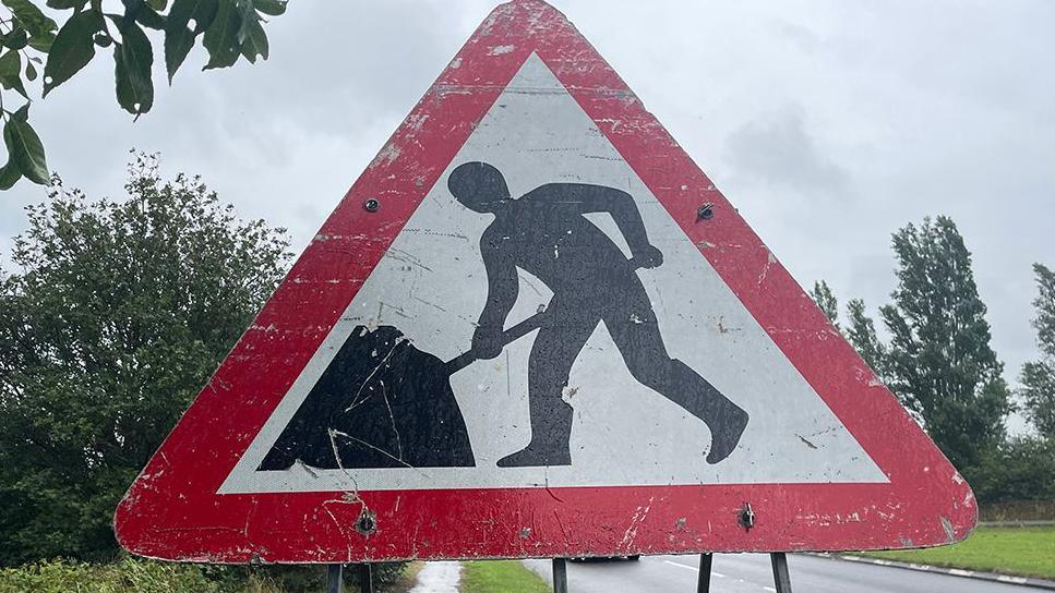 A close up of a triangular roadworks sign, showing a silhouette digging in a red triangle. A road and trees can be seen in the background