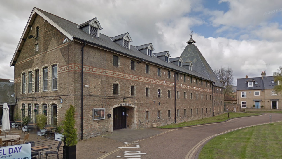 A long building, which is three stories, goes across the whole length of the image. The brown brick building has lots of rows of small windows and an arch entrance with a door on the left hand side. 