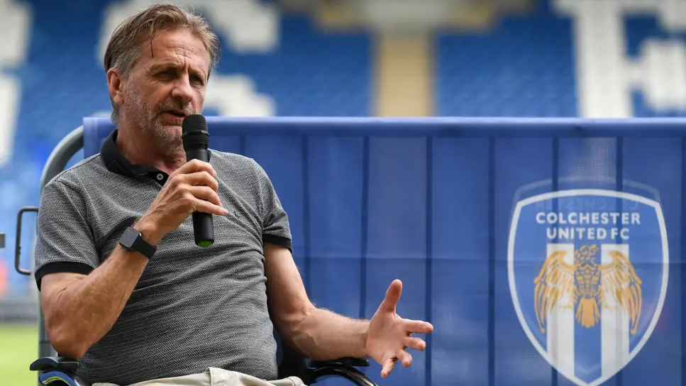 A mid shot of Robbie Cowling. He has greying hair and is sitting in a chair speaking into a microphone. Behind him is a fence panel with Colchester's logo and branding draped over it.