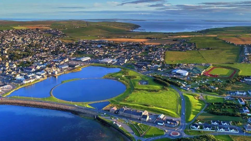 An aerial view of Orkney by the sea which shows a green landscape with homes and buildings connected by roads, large blue lakes and a running track.