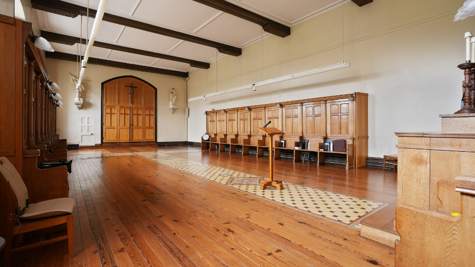 Inside one of the rooms. It has cream walls and brown wooden beams across a white ceiling. In the middle of the room is a stand with a microphone and what looks like an altar at the front with individual prayer stools down the side. At the back of the room are two large wooden doors with a cross on the top.