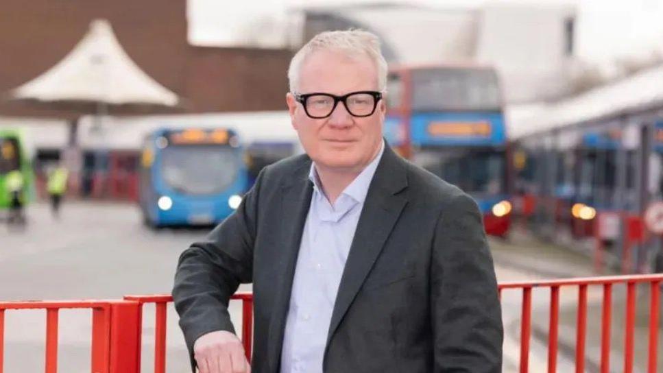 Richard Parker wearing black glasses, a grey blazer and a light blue shirt. He is standing in front of buses at a depot