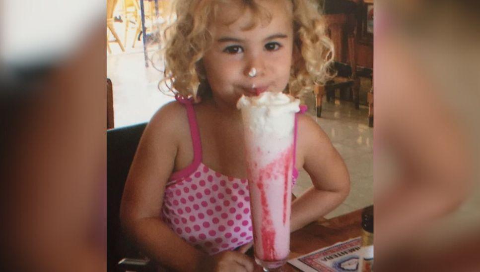 A child in a pink bathing suit is seated in a restaurant. She is drinking a strawberry drink with whipped cream on top. 