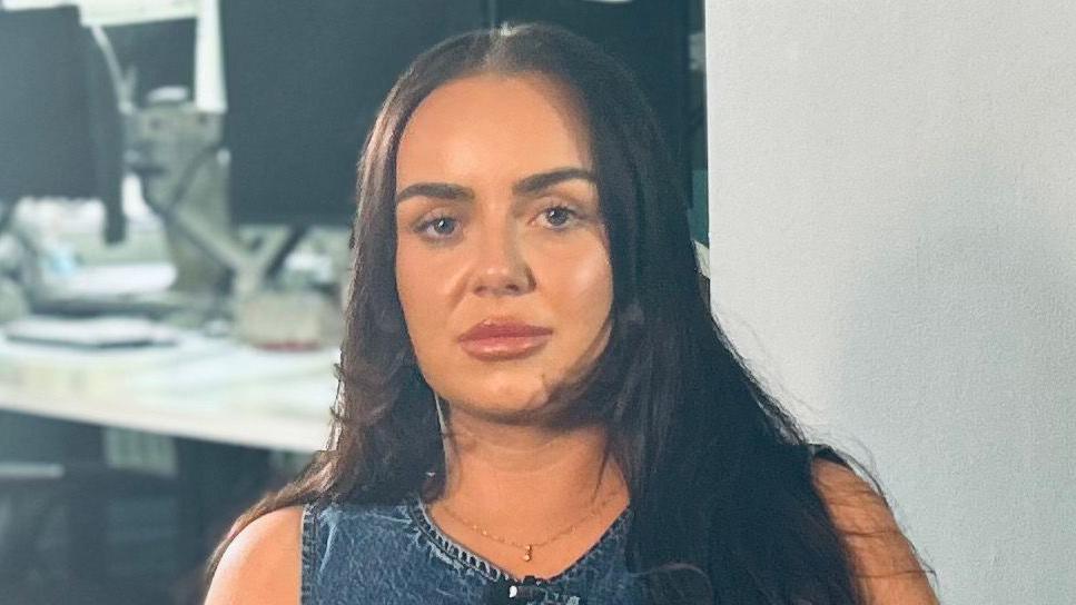 Haileigh has long brown hair. She is sitting in an office and looking directly into the camera. She's wearing a gold necklace and a denim top.