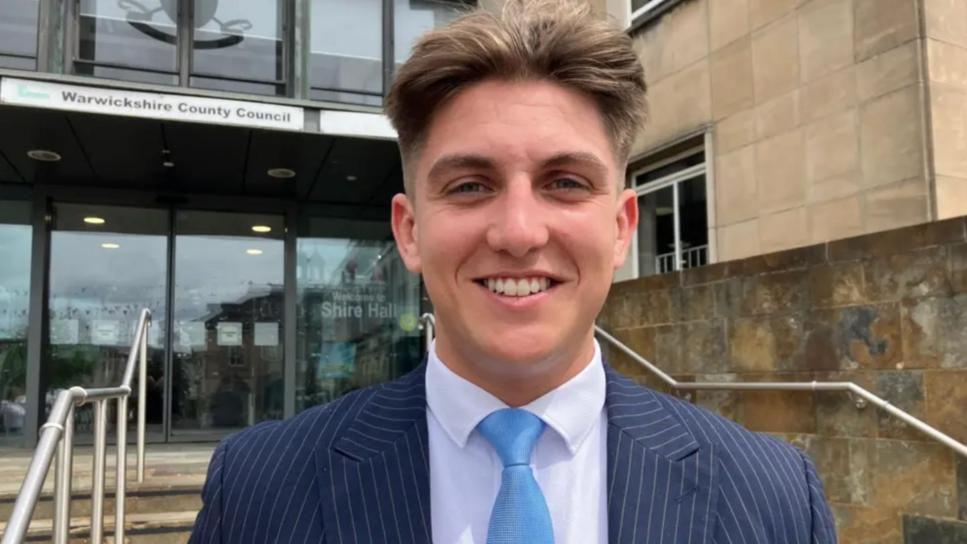A young man with short light brown hair is wearing a blue pinstriped suit, a white shirt and pale blue tie. In the background are steps leading up to a large glass building with a sign above double doors that reads, "Warwickshire County Council".