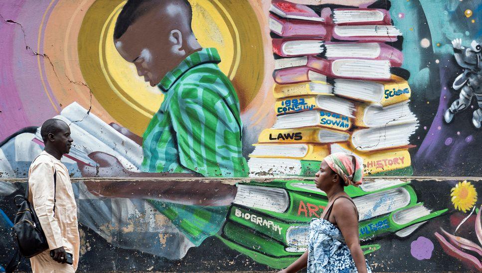 People walk past a mural outside a school building in Accra showing a boy reading a book as he leans against a big pile of books.