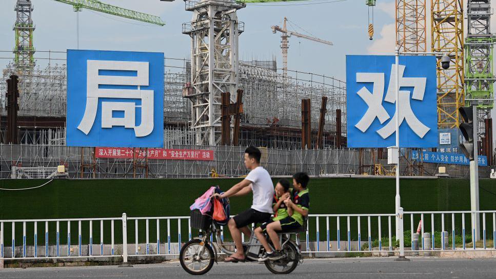 People commute in front of the under-construction Guangzhou Evergrande football stadium in Guangzhou, China's southern Guangdong province on September 17, 2021. The photo shows a male motorcyclist with two passengers, children, seated behind him, as they ride past a large construction site.