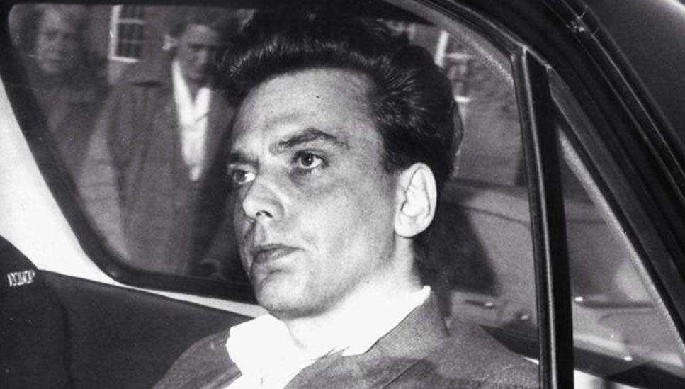 A black and white photograph showing the Moors murderer Ian Brady, seated in the back seat of a car. He has dark hair and is wearing a suit jacket. The car window is partially visible, with another individual standing outside the vehicle, looking in.