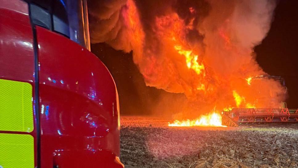 A field with a fully alight combine harvester in field of stubble. It is covered in orange flames and smoke, which is rising up into the black night time sky. On the left is the corner of a fire engine and on the right a firefighter can just be glimpsed.