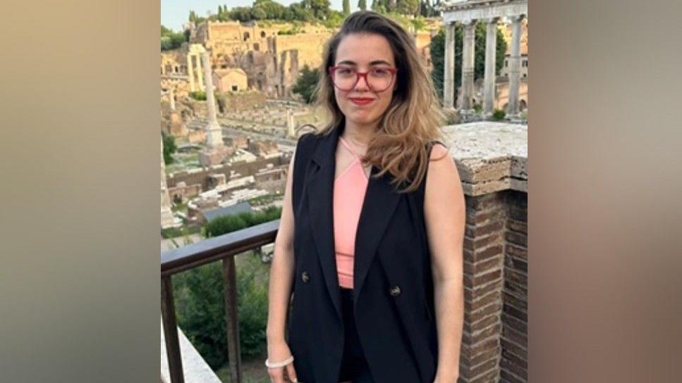 Laura Carrillo, wearing red glasses, brown hair to the shoulders, a black jacket and pink top standing next to a wall