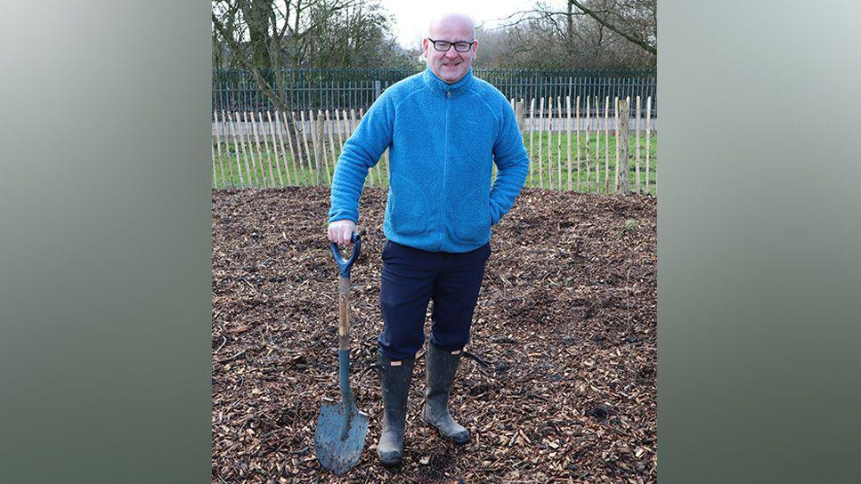 Councillor Shaun Turner wearing a blue fleece and navy trousers and wellies holding a spade resting on the ground in a park where new trees are being planted