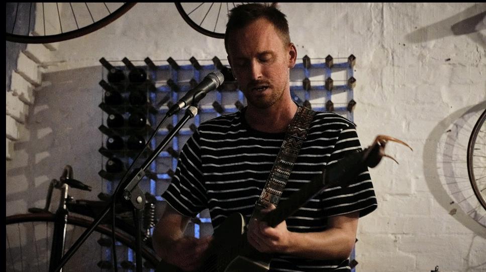 Tom wears a black t-shirt with white stripes. He plays guitar on stage, standing in front of a microphone. A white brick wall is behind him with vintage bikes on display. He has closely-cropped light hair and fair facial stubble.