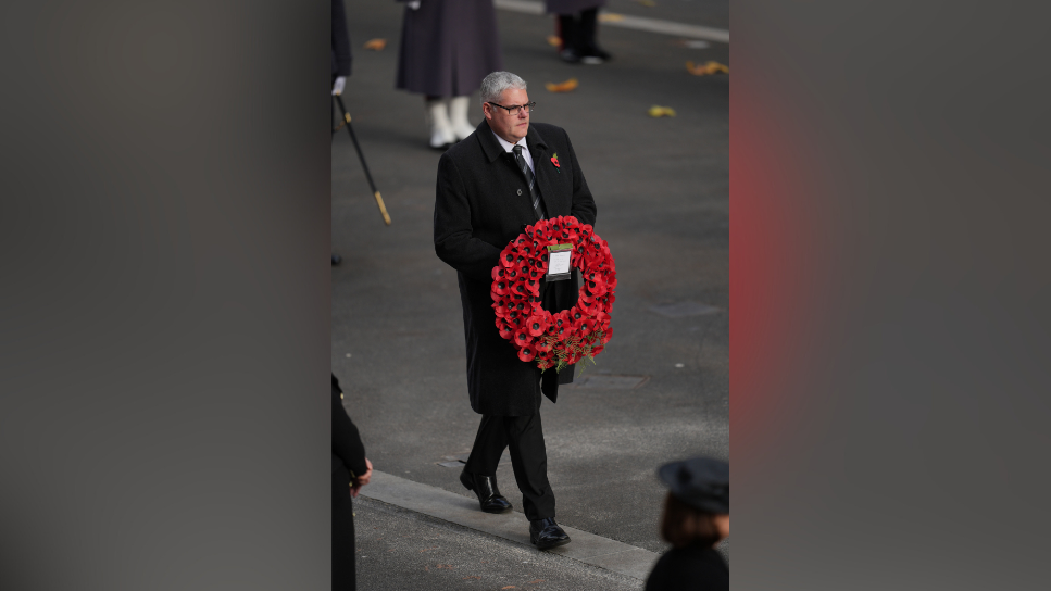 Gavin Robinson is holding a wreath made out of poppies. He's dresses in black and walking through a road with other people.