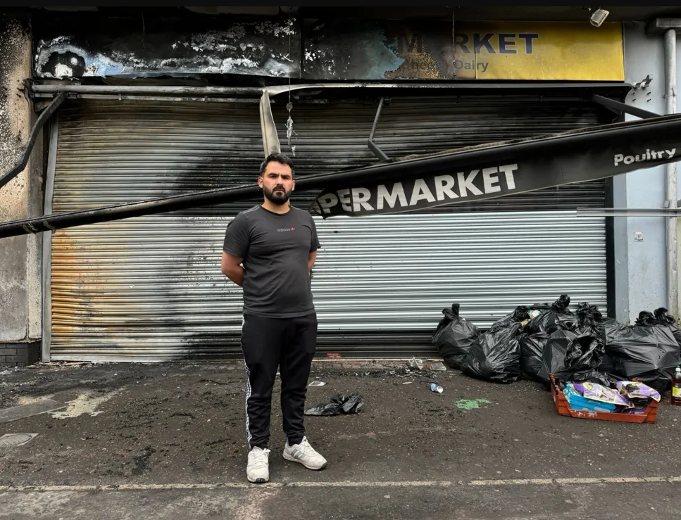 Abdelkader standing in front of his store where the signage has been destroyed by fire and bags of rubbish are visible in front