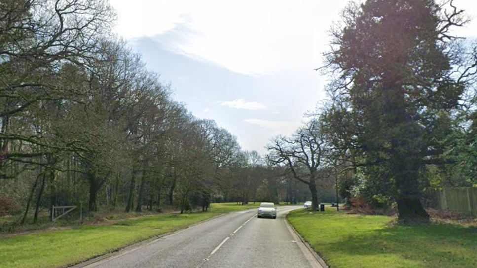 A single carriageway road, with woodland to the left, and some trees and a wooden fence to the right. A silver car is driving towards the camera. There is a blue sky and cloud above.