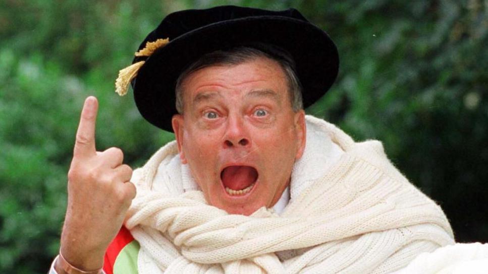 Dickie Bird: a man wearing a black hat with a yellow tassel and a large white scarf around their neck has their mouth open and eyes wide, with one hand raised and pointing upward. The background features blurred greenery.