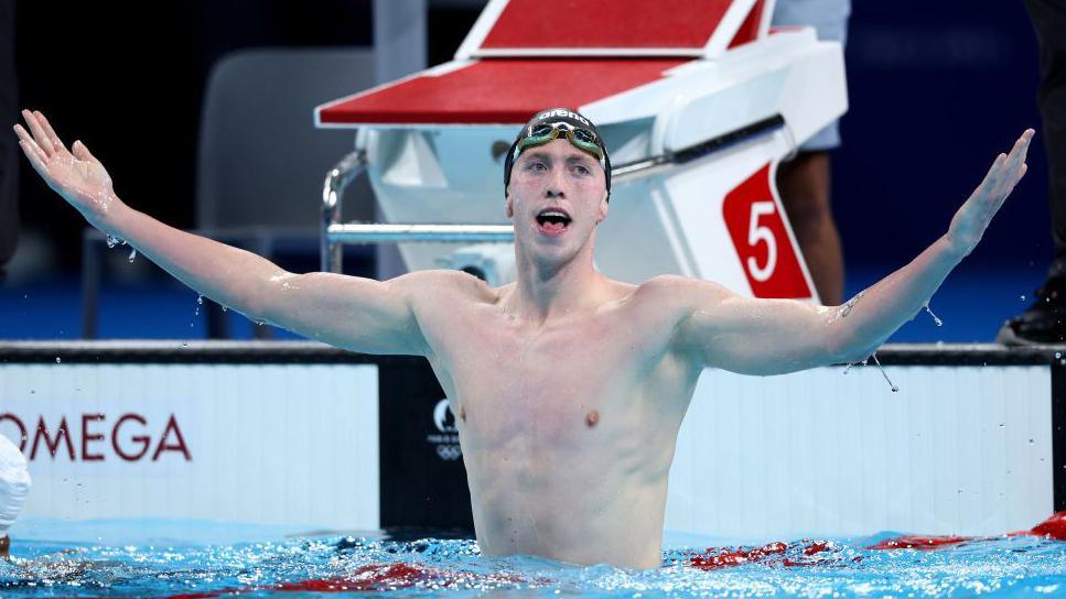 A swimmer raises his hand up in the pool, he has on a black swim cap and green goggles