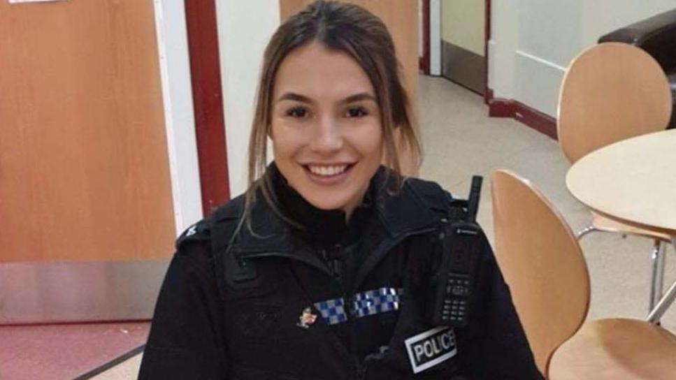 PC Molly Bury sits at a classroom in police uniform, she is smiling nd has dark hair tied up at the back