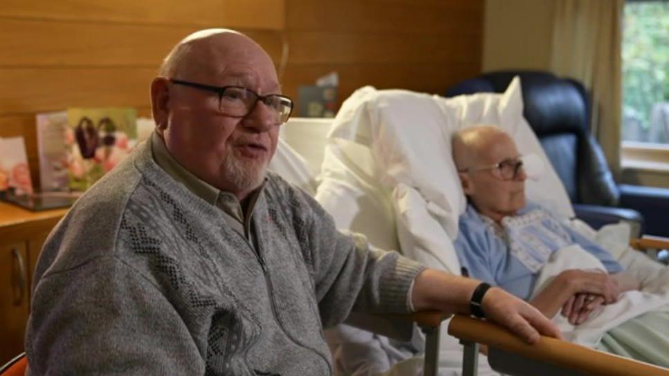 A man looks at the camera and speaks to the reporter with his hand on the arm of a chair. Behind him his wife lies in a hospice bed wearing glasses and blue night clothes looking straight ahead. 