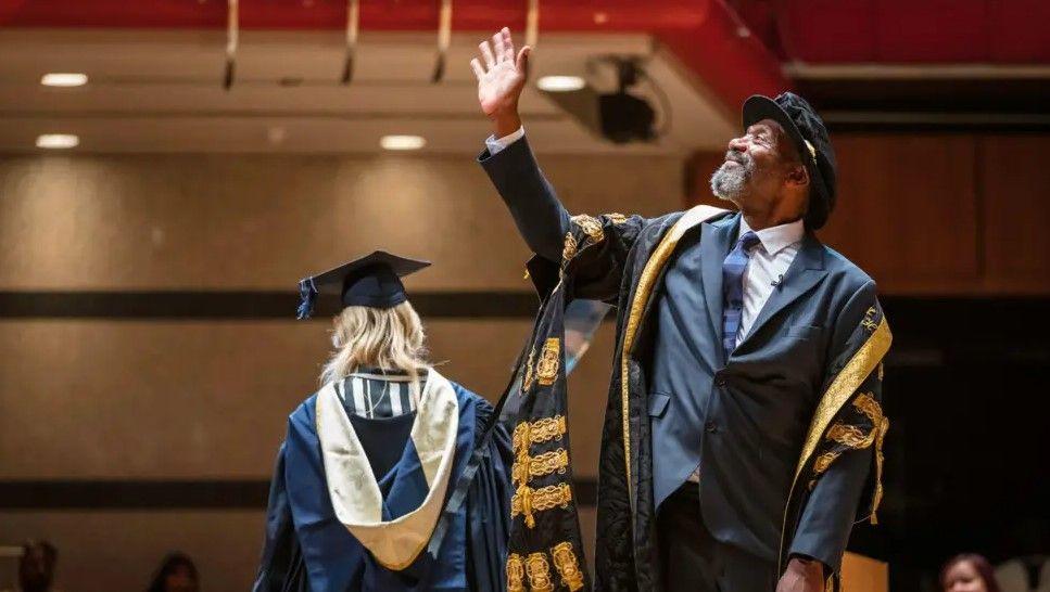 A man wearing a dark grey suit with a navy tie under black and yellow formal academic gowns. He is waving. Behind him a woman wearing similar gowns has her back facing the camera