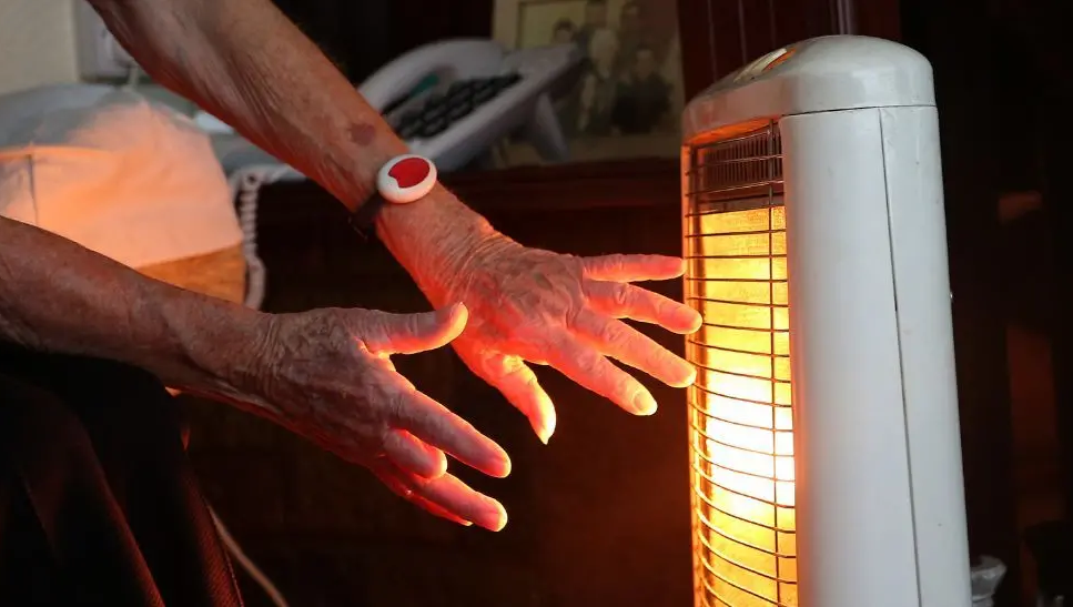 An older person warms their hands at a heater