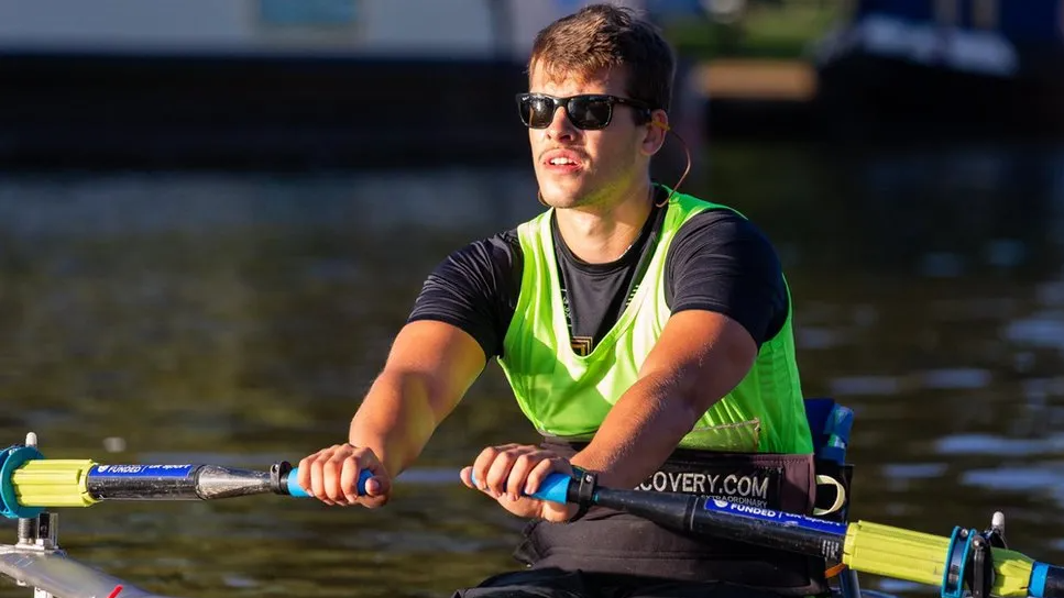 Xander Van der Poll rowing in a river, wearing a black shirt, green sports bib, and black sunglasses