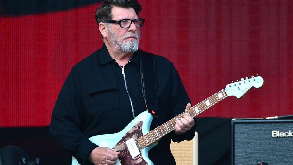 Will Sergeant standing on stage in front of a red background playing the guitar. He is wearing glasses and standing beside a speaker. 