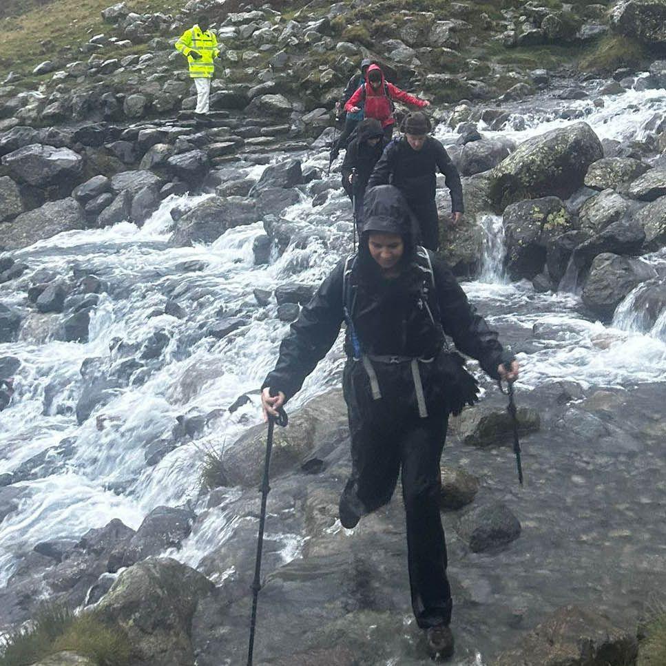 Six hikers crossing the flooded path. They are wearing waterproof coats and hiking bags. A woman leads the group at the front and is holding a walking pole in both hand. The stone steps of the path lead down the hill towards the camera. A river is flowing downhill from the right straight across the path. It has flooded out of its usual waterway and is covering much of the path.