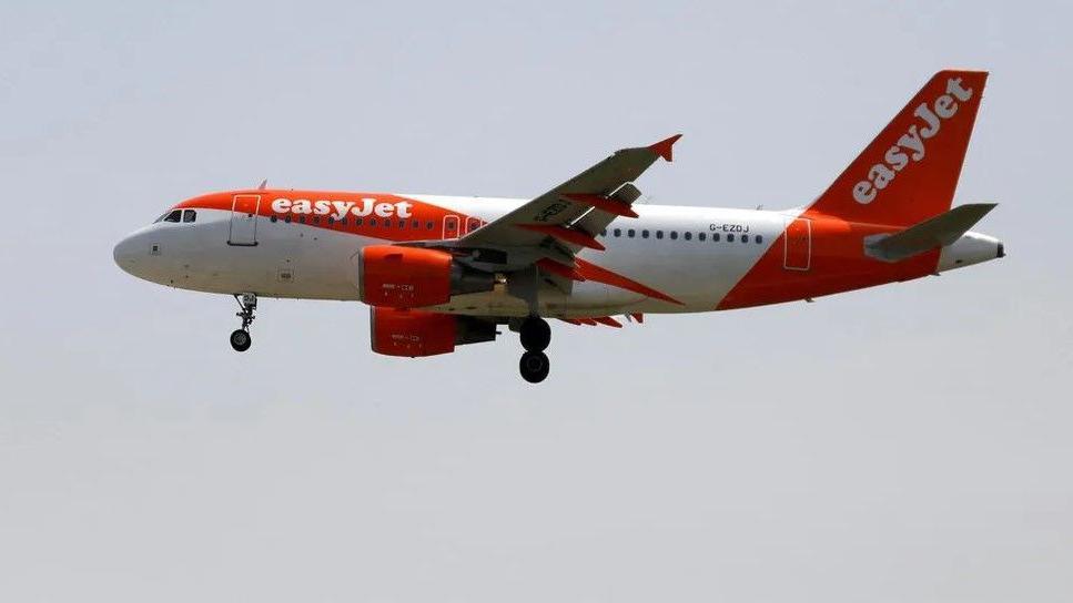 An easyJet-branded plane, with orange and white livery, is seen in flight against a light grey sky