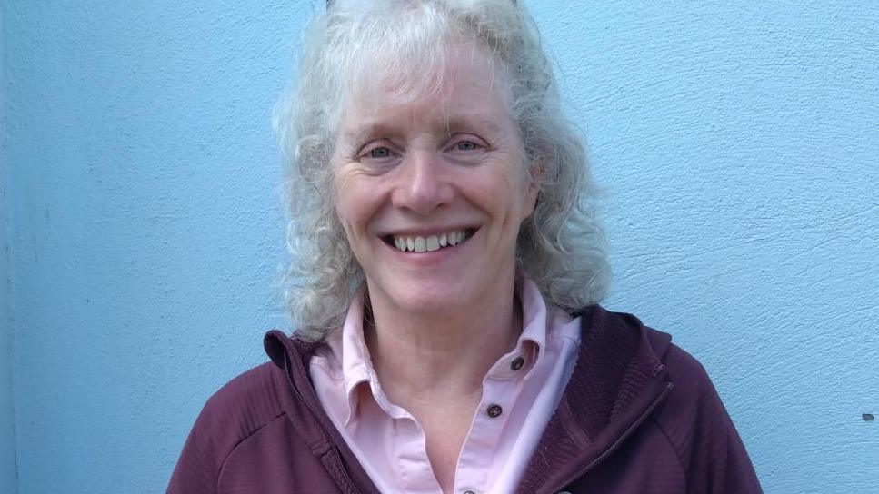 Pam Ashcroft, a woman with curly grey hair and a fringe smiles, she wears a pink shirt and stands in front of a blue wall.
